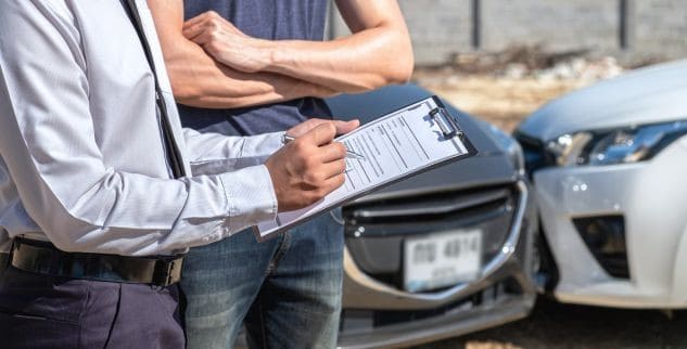 Two men stand by cars involved in an accident, with one man writing on a clipboard.