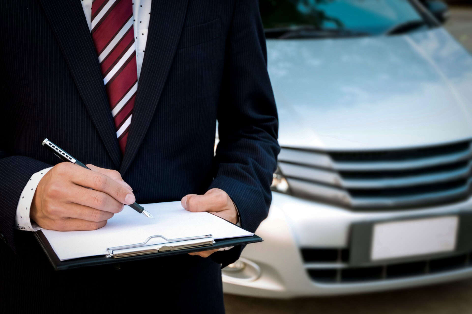 A person in a suit holding a clipboard and pen stands in front of a silver car.
