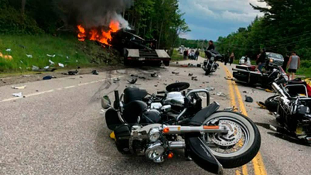 Motorcycle accident scene on a road with debris and a vehicle on fire in the background. People are present and observing the situation.