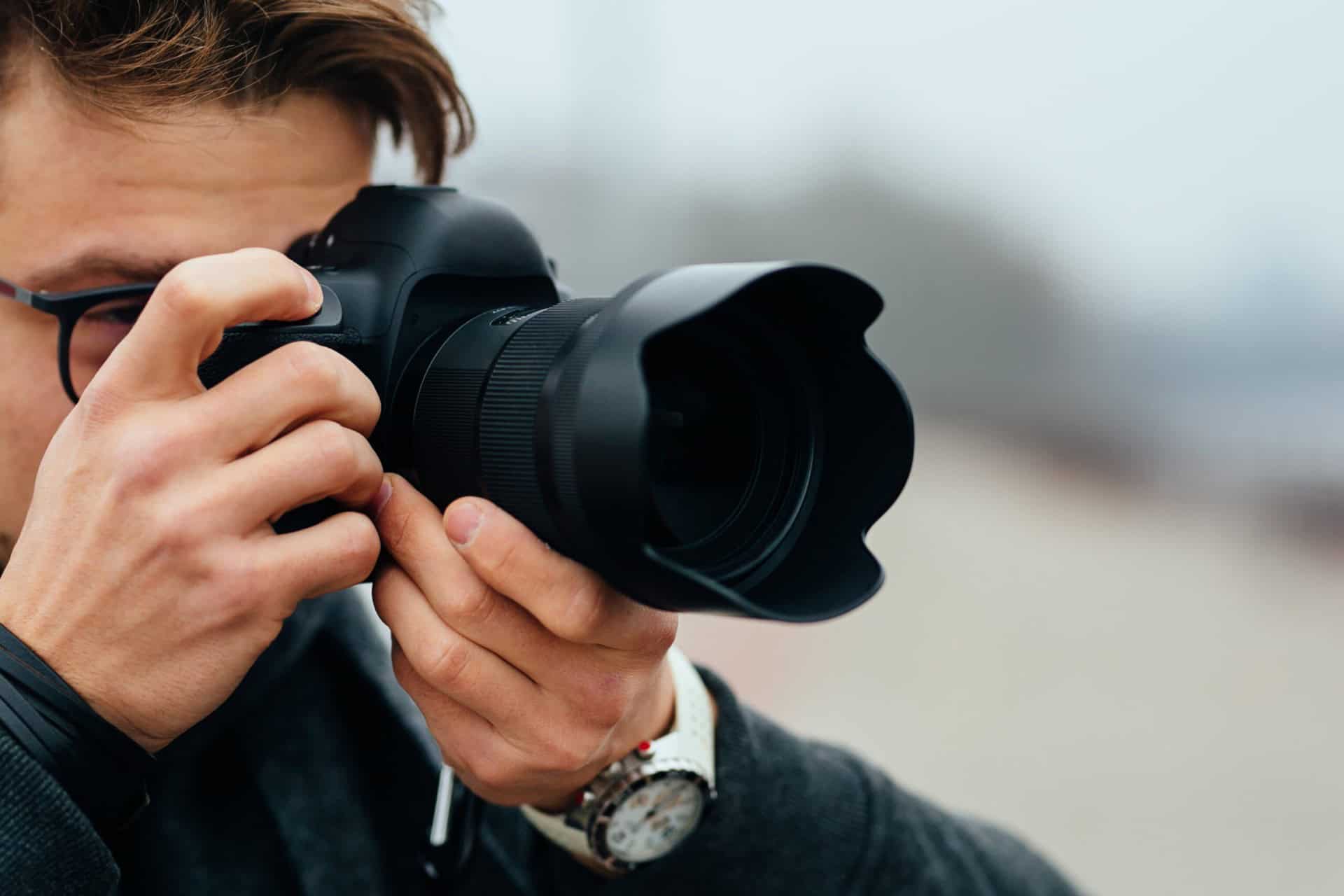 A person with glasses holds a camera close to their face, focusing on taking a photograph outdoors.
