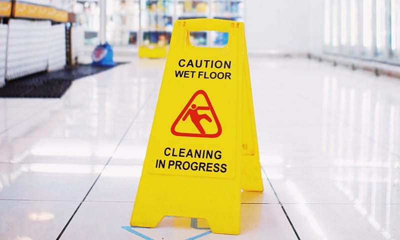 Yellow caution sign on white tiled floor reads "Caution Wet Floor Cleaning in Progress" with a slipping figure icon.