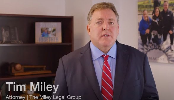 Man in a suit and red tie stands indoors with bookshelves and a family photo in the background. Text reads: "Tim Miley, Attorney | The Miley Legal Group.