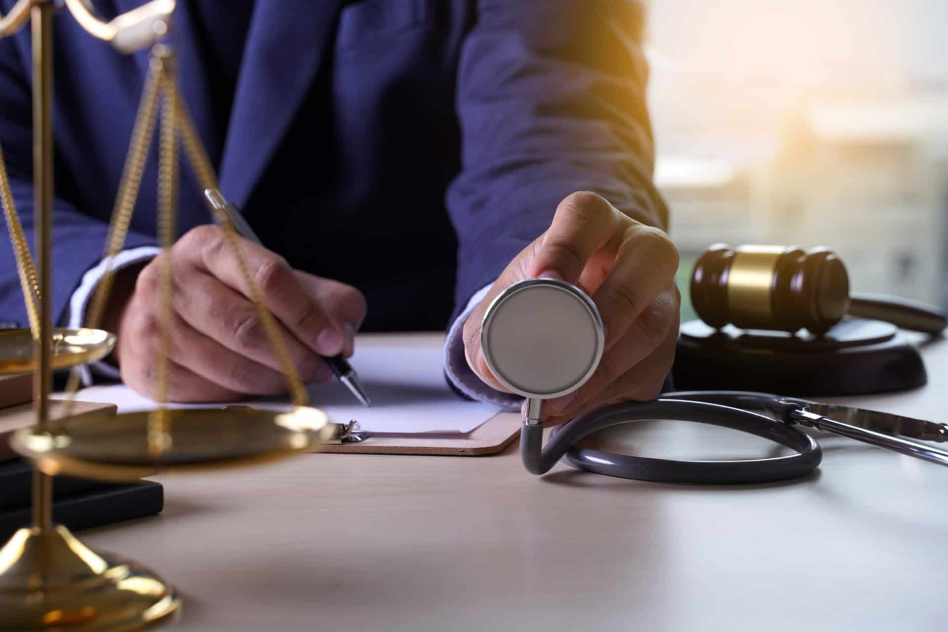 A person holds a stethoscope in one hand while writing on a notepad at a desk with a gavel and scales of justice nearby.