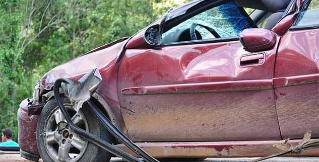 Damaged red car with dented body and broken windshield in a wooded area.
