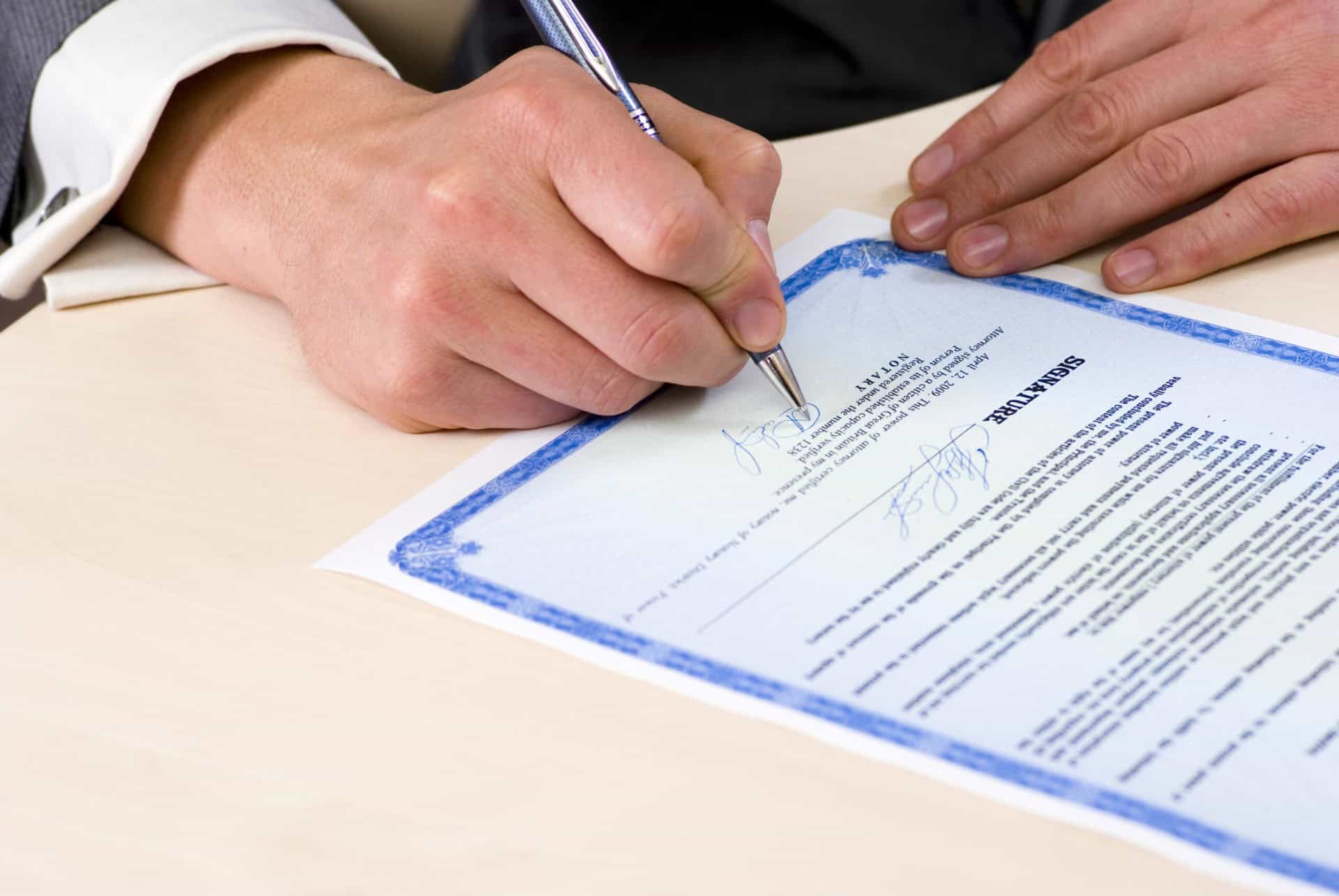 A person in a suit signs a blue-bordered document on a desk with a pen.