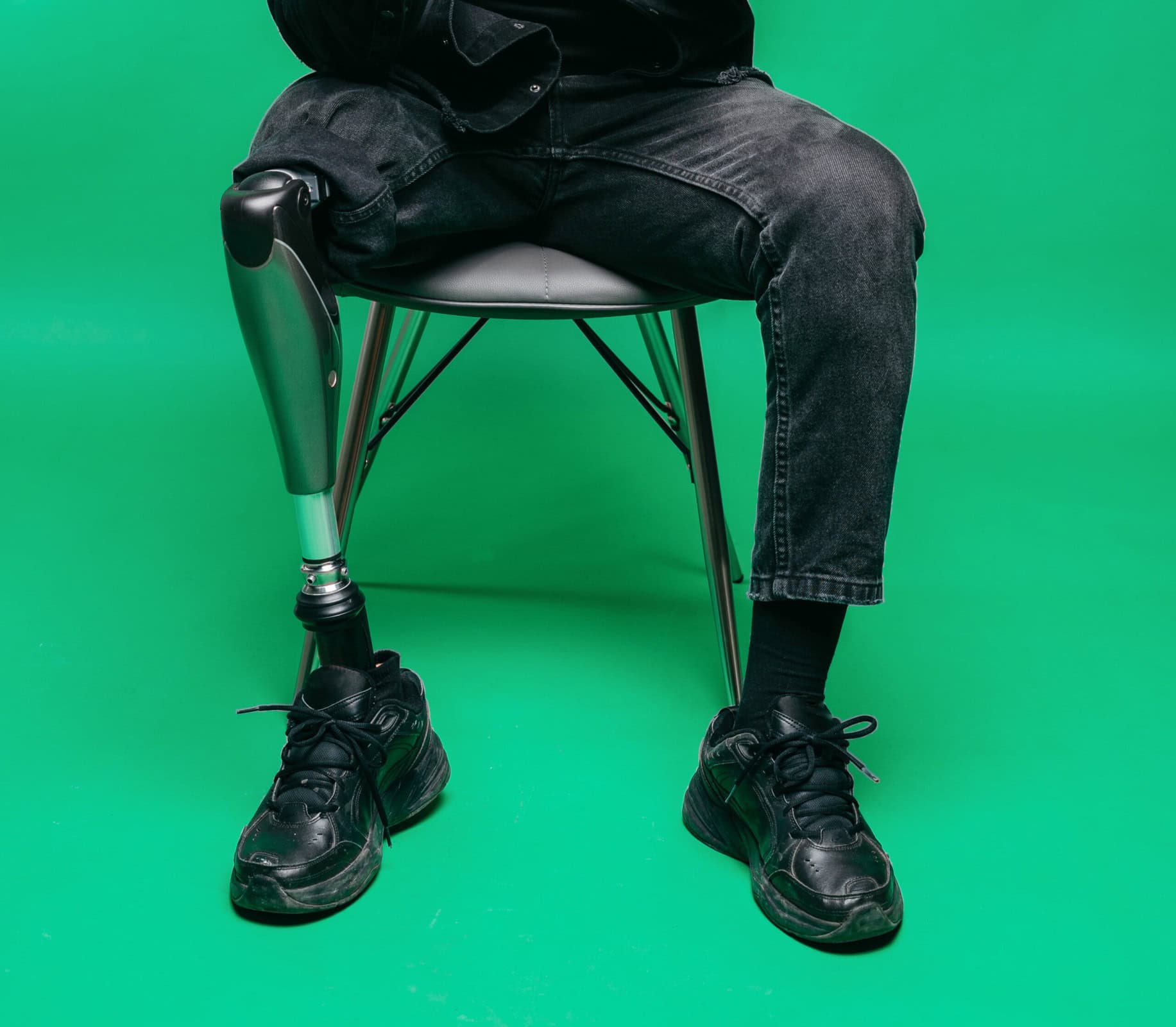 A person sitting on a chair with a prosthetic leg, wearing black pants and shoes, against a green background.