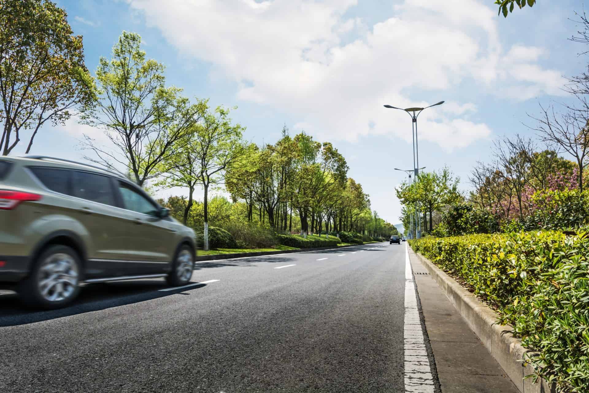 A car drives on a tree-lined road under a partly cloudy sky.