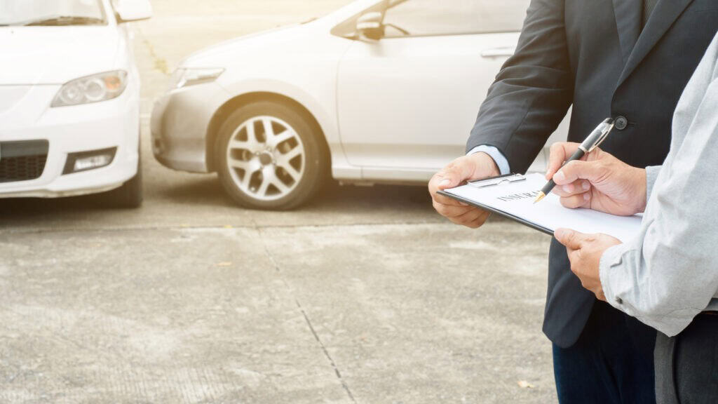 Two people in formal attire signing documents on a clipboard, standing outdoors. Two white cars are parked in the background.