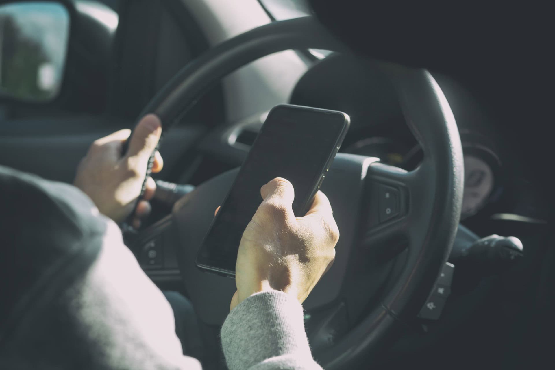 Person driving a car while holding and looking at a smartphone in their right hand, with both hands on the steering wheel.