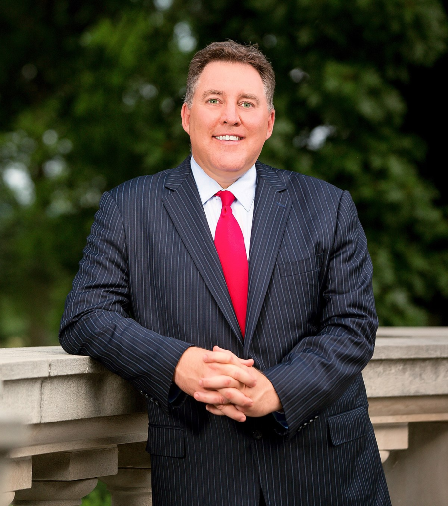 Man in a pinstripe suit with a red tie stands outdoors, leaning on a stone railing, with trees in the background.