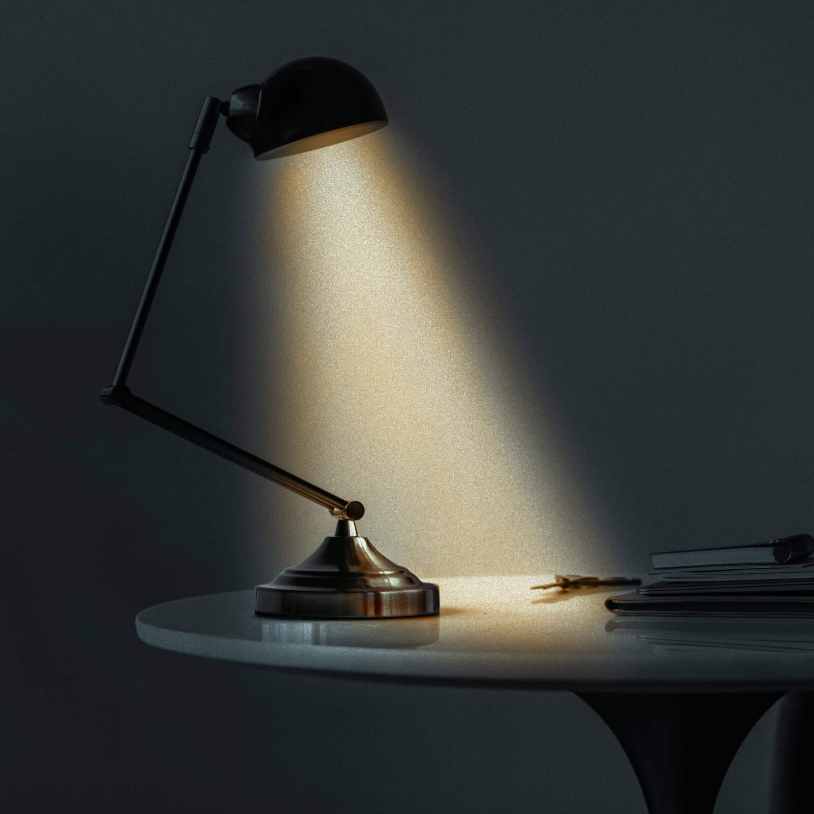 A black desk lamp illuminates a small area of a round white table, where there are books and a pair of glasses in a dimly lit room.