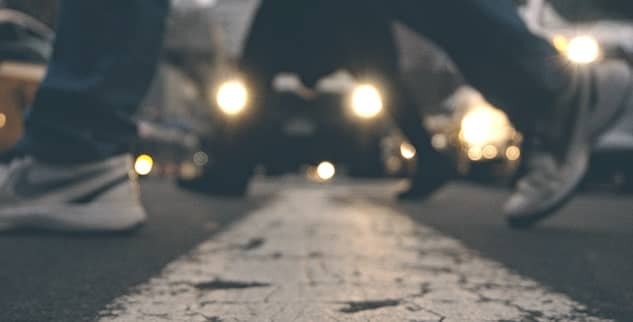 Blurred image of people walking across a street with a white line in the foreground. Car headlights are visible in the background.
