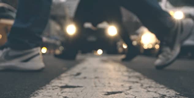 Blurry image of pedestrians crossing a street with cars and headlights in the background.