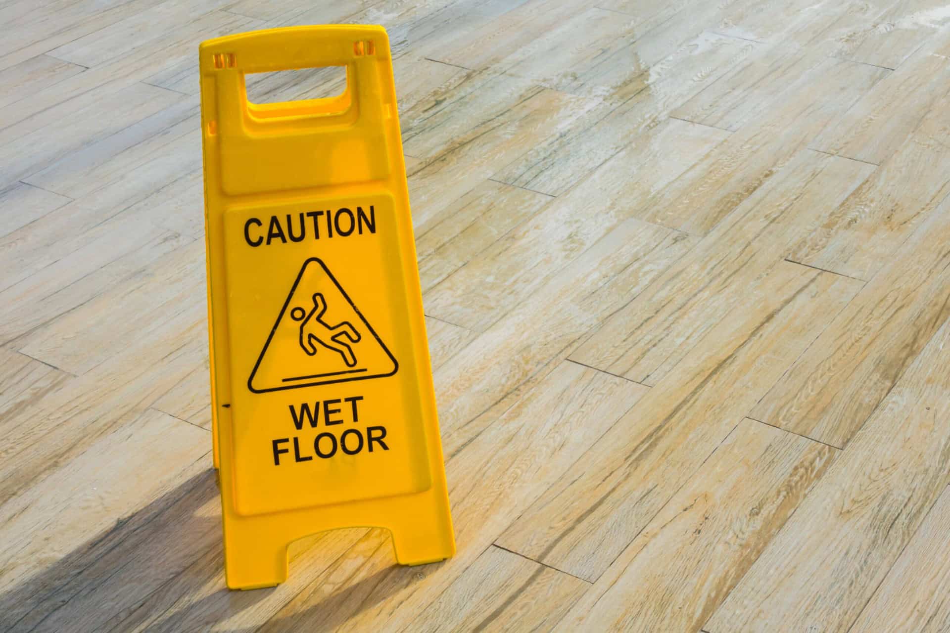Yellow caution sign on wooden floor reads "Caution Wet Floor" with a slipping figure icon, indicating a potential slip hazard.