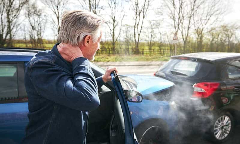An older man with neck pain stands by an open car door. Two cars have collided, with visible damage and smoke coming from the engine of the front car.