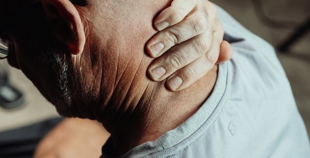 A person with short hair and glasses rubs their neck with their left hand, wearing a gray shirt.