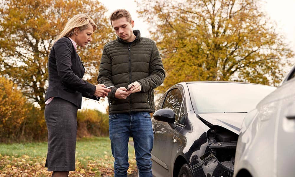 Two people exchanging insurance information after a car accident. Both examine documents by a damaged vehicle on a rural road with autumn trees in the background.