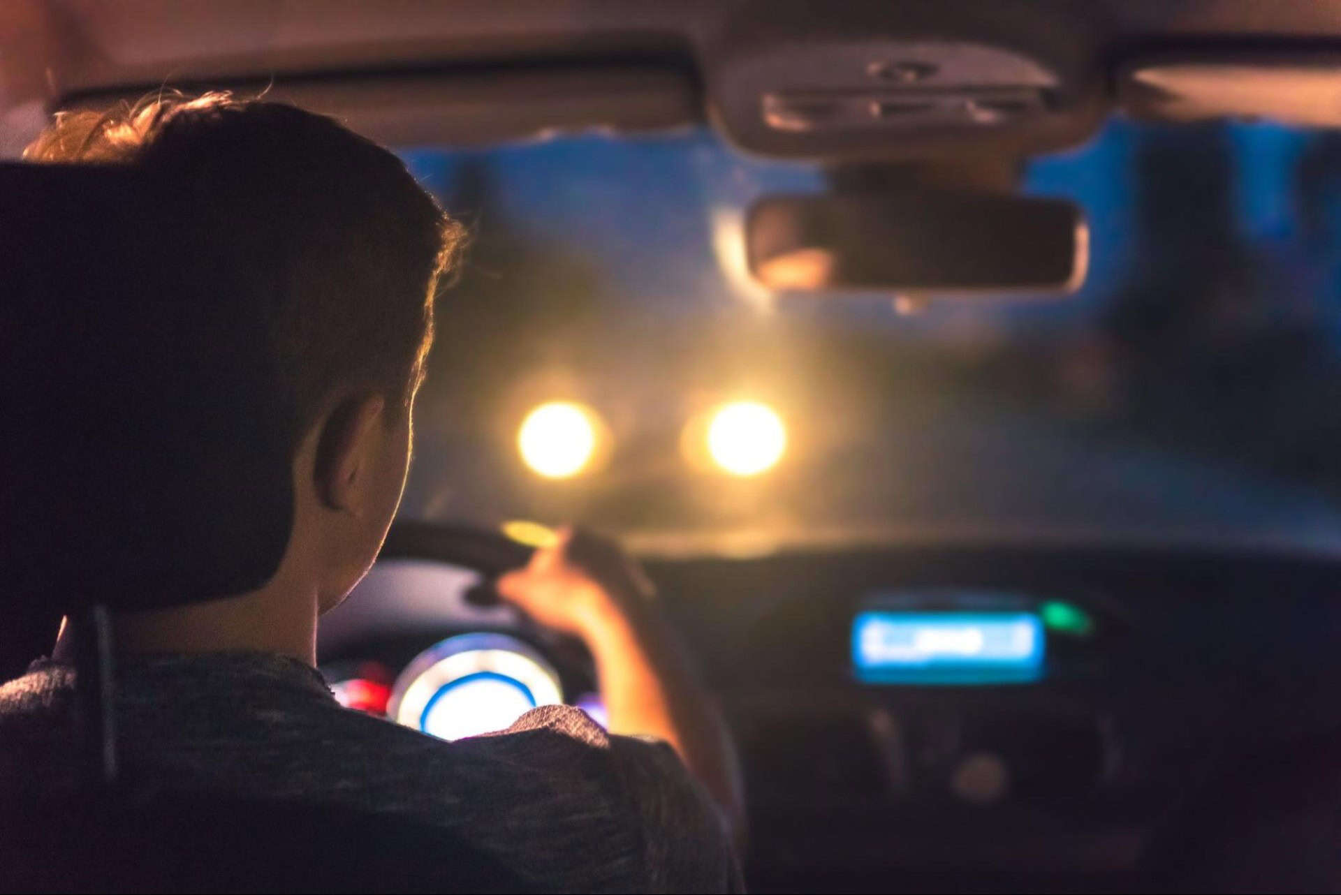 Person driving a car at night, illuminated by oncoming headlights. Dashboard lights are visible.
